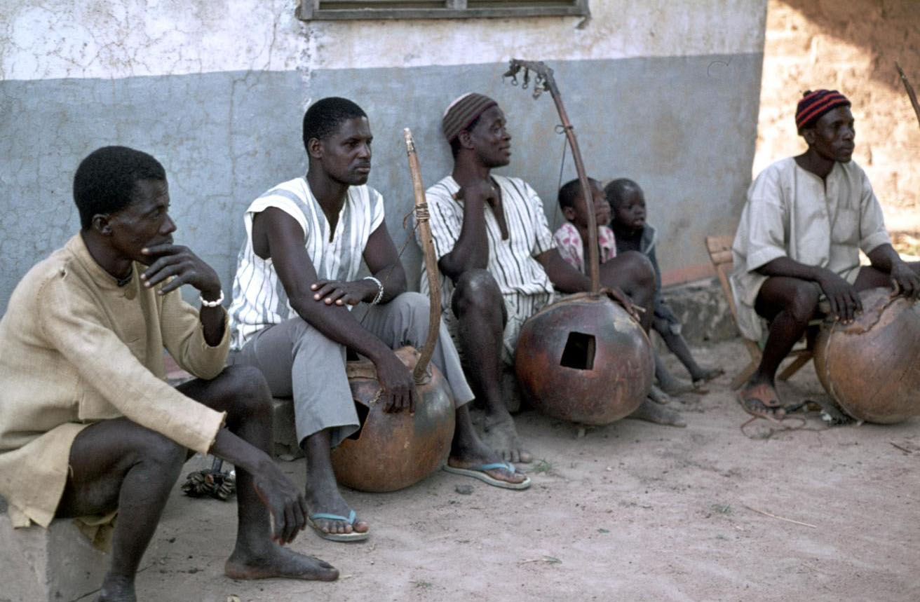 Slide of three men playing the balogo or harp, another man and two children sit next to them: Kakoulogo, Ivory Coast, 1977, photographed by Jean Jenkins.