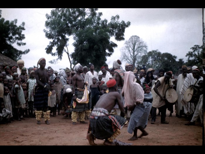 Slide of dancers wearing leg rattles and a dancer with a baby, two men accompany them on the drums: Shaki, Nigeria, 1960, photographed by Jean Jenkins.