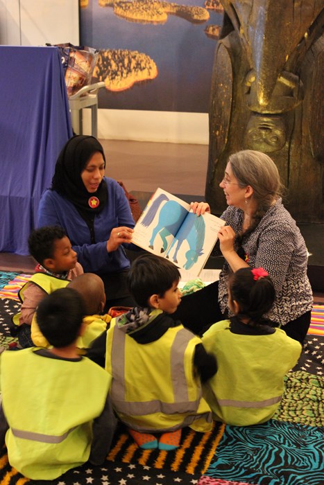 Two women read a story and show the book to a group of children sitting on the floor
