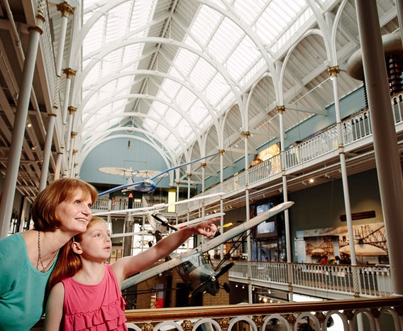 A woman and child stand on the balcony in front of different model airplanes hanging from the ceiling. The child points to something out of frame.