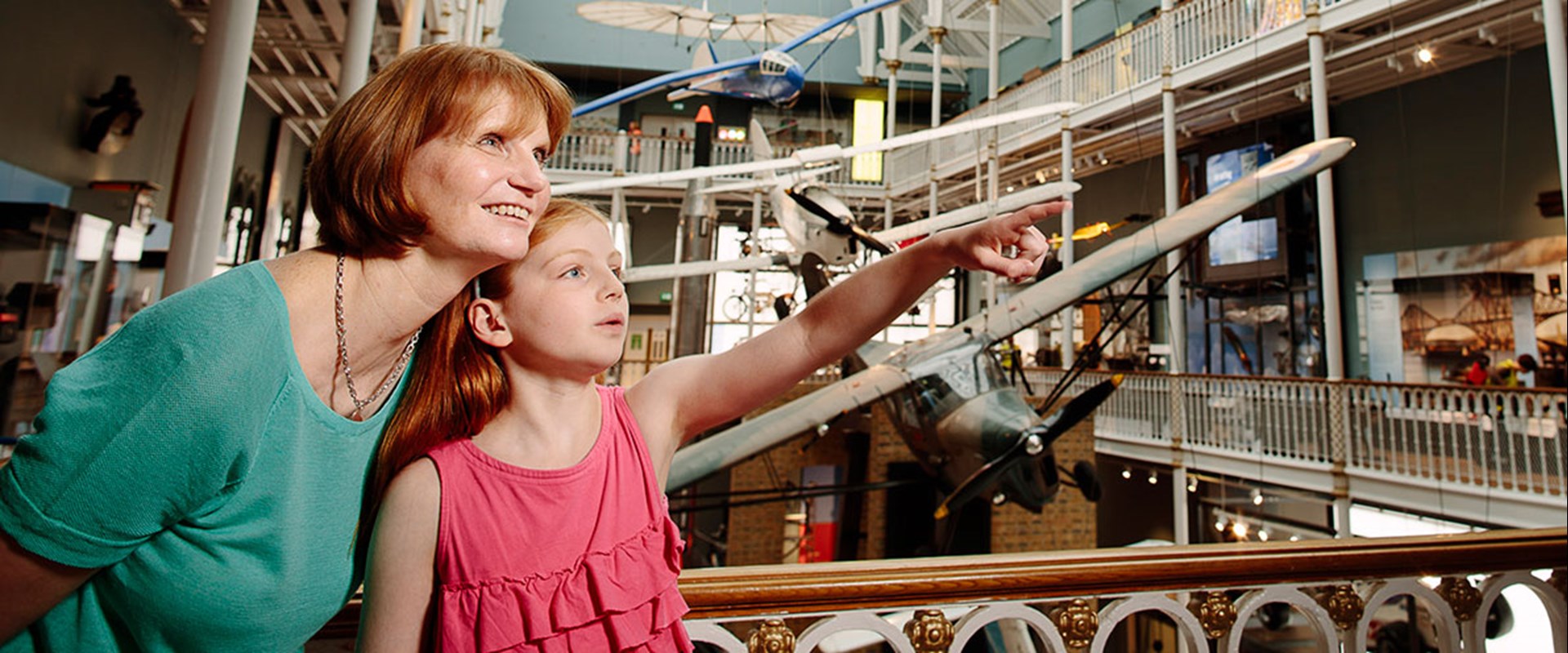 A woman and child stand on the balcony in front of different model airplanes hanging from the ceiling. The child points to something out of frame.