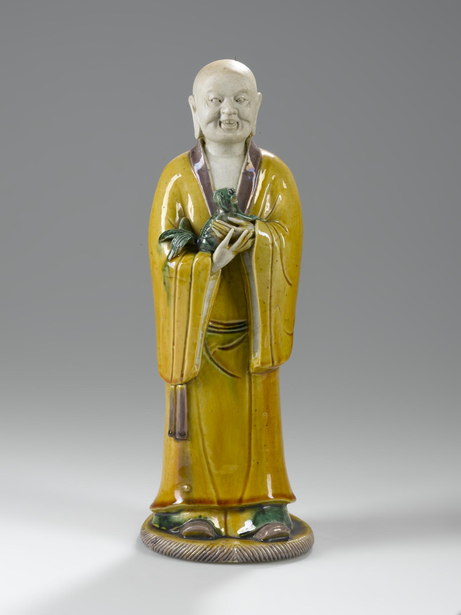 Figure of a luohan or Arhat, holding a lion cub in his arms in porcelain with sancai (three coloured) glazes, possibly Xiaoshi Luohan (Luohan with laughing Lion): China, Ming or Qing Dynasty, 16th - 18th century AD. 