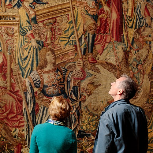 Two visitors look at a large tapestry hanging on a wall.