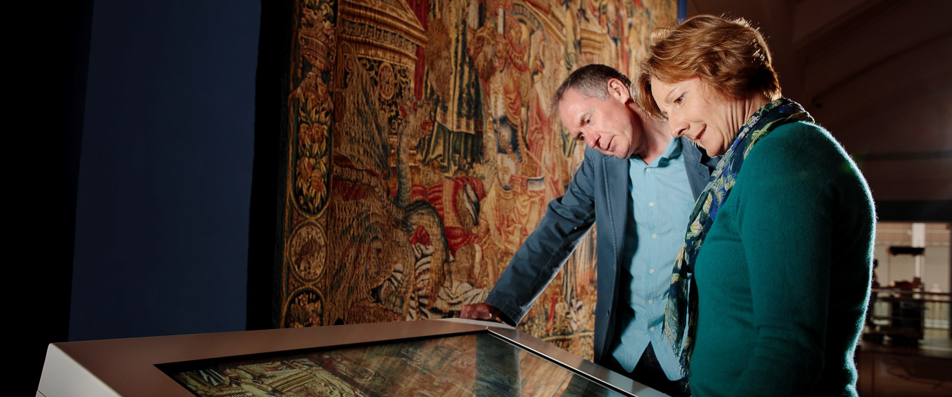 Two visitors looking at an interactive touch screen in front of a large tapestry.