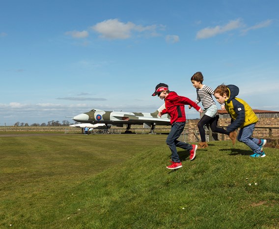 Three children running down a hill with a large aeroplane in the background.