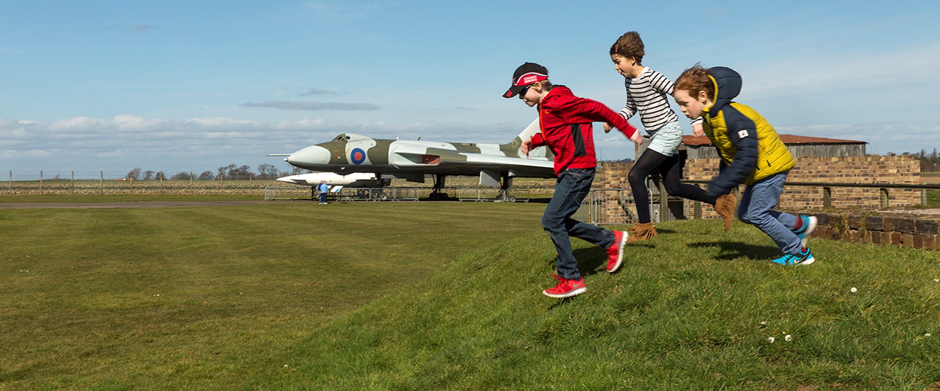 Three children running down a hill with a large aeroplane in the background.