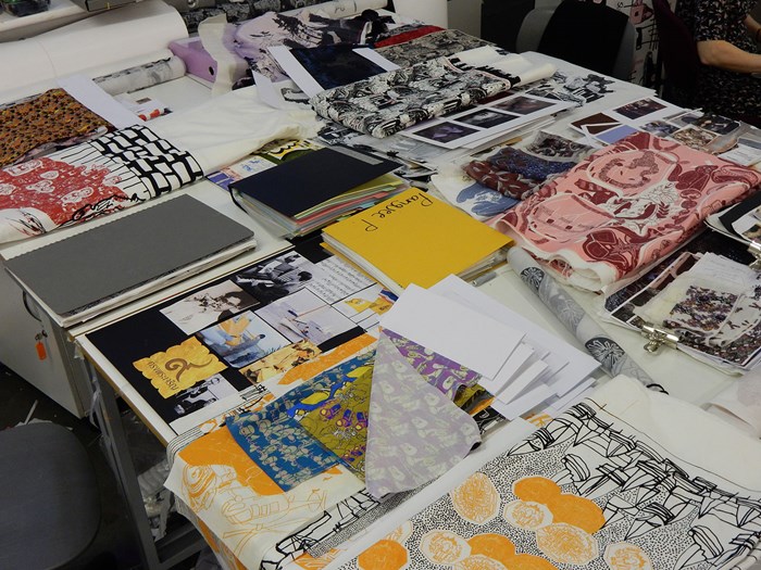 A selection of colourful student-designed cloths folded with other images and papers on a table