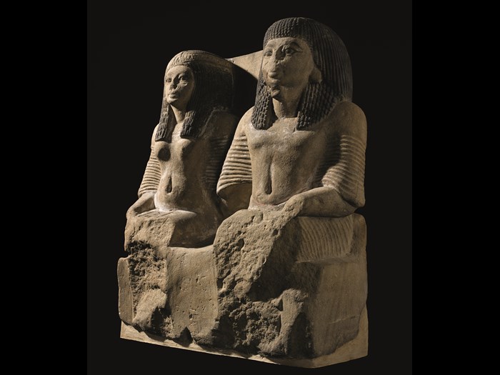 Pair statue in fine yellow sandstone of a Chief of the Police and his wife seated side by side, wearing long pleated robes, collars and heavy wigs: Ancient Egyptian, excavated at Sheikh Abd el-Qurna, Thebes, 19th Dynasty, c.1291-1188BC.