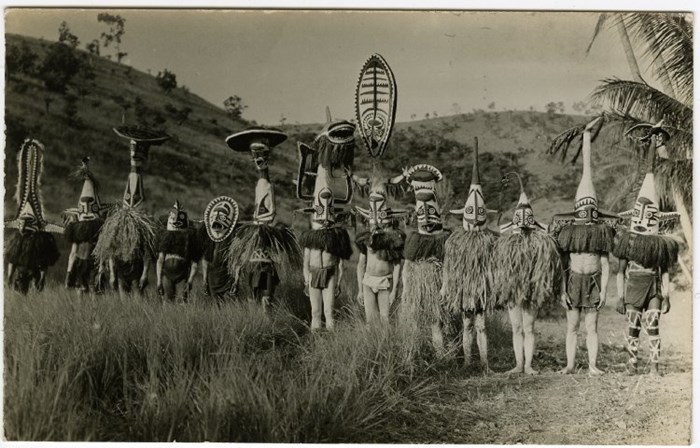 Postcard showing thirteen young men outdoors wearing eharo masks; they also wear plant fibre shoulder coverings and cloth wraps, as well as having thier bodies painted; grass and hills behind them; Purari Delta; Papua New Guinea. © The Trustees of the British Museum (CC BY-NC-SA 4.0)