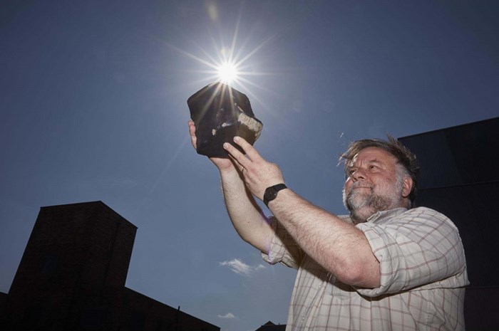 Peter Davidson, Senior Curator of Mineralogy, holds a fragment of the Strathmore Meteorite against a clear sky with the sun in the centre of the frame