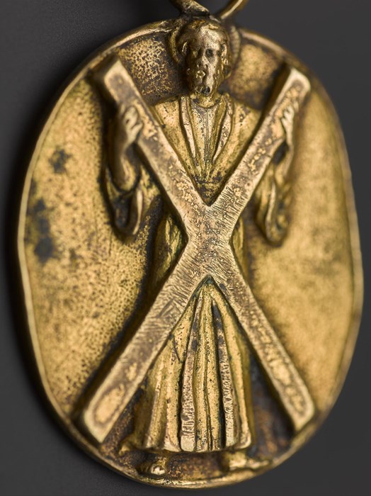 Oval badge of gilt brass, featuring images of a thistle head and the figure of St Andrew, associated with the Order of the Thistle. Made in the late 17th – early 18th century.