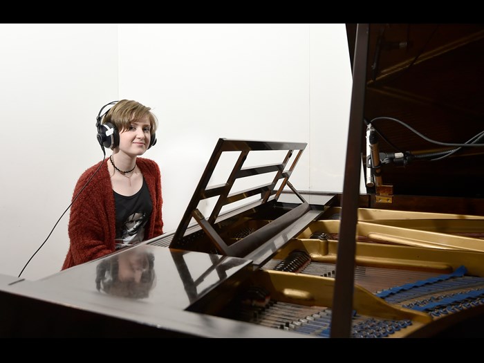 A girl with short hair and a red cardigan wears large, over-ear headphones and sits at a partially-visible grand piano with the top open.