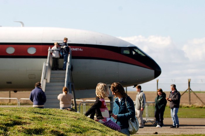 A child sitting on a small hill. A larger group of people are in the distance standing outside of a Comet aeroplane.
