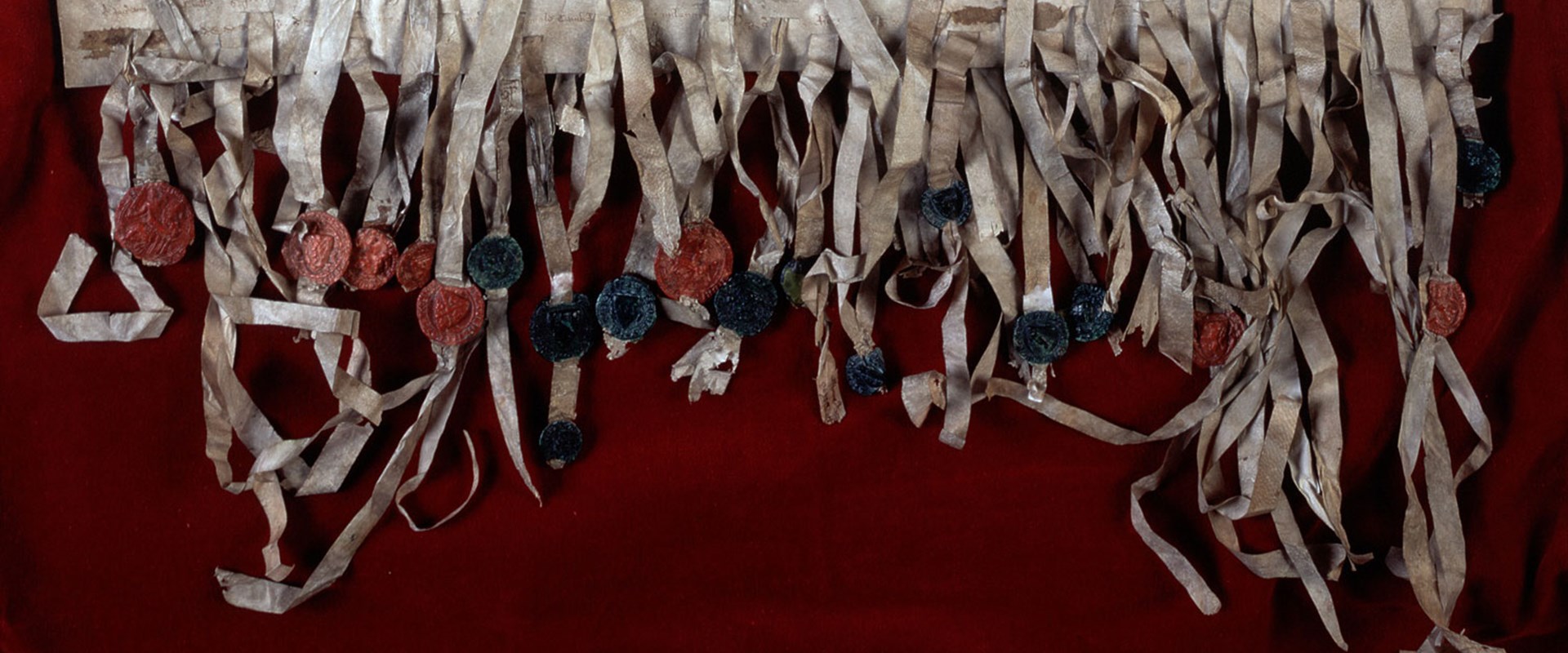 Wide, rectangular crop of the bottom section of the Declaration of Arbroath, a weathered medieval document on a dark red surface. Dozens of paper tassels are affixed with red and green wax seals.