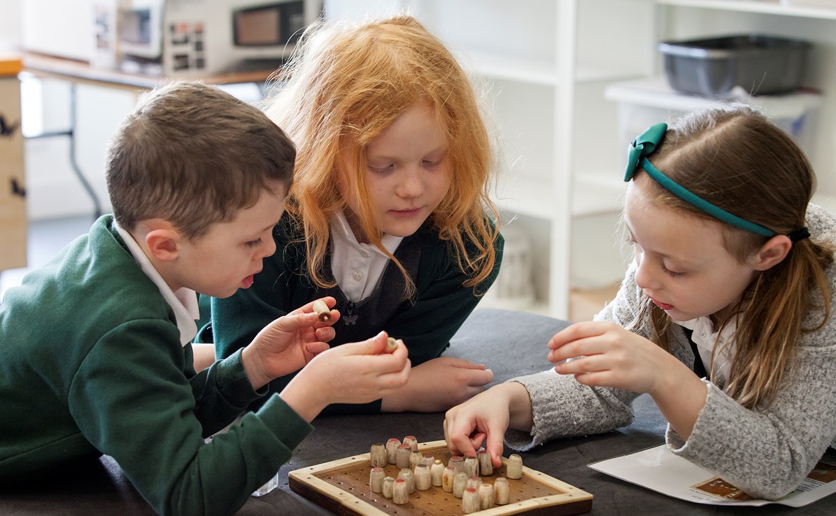 Three primary school pupils are gathered around and are playing a board game. 
