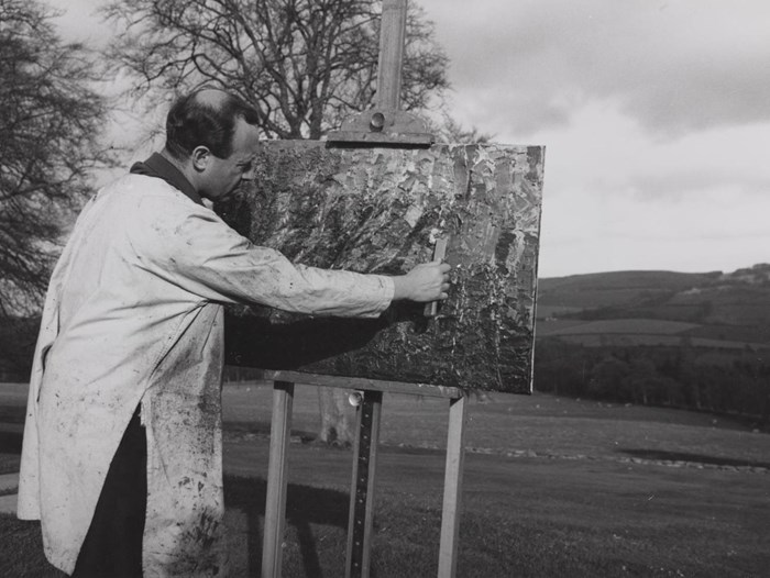 Bernat Klein painting outside, at an easel with a palette knife, wearing a white smock.