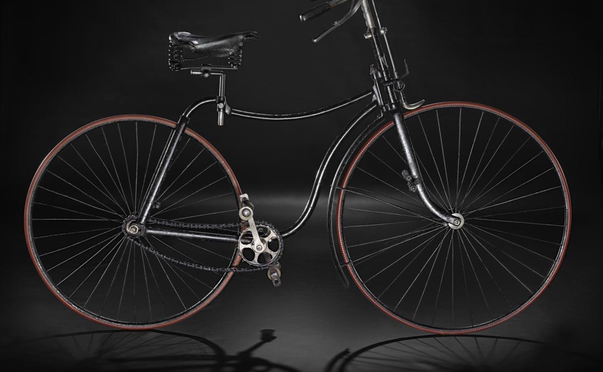 A vintage black bicycle with a light brown rim around the wheels, viewed from the side and standing upright against a black background.