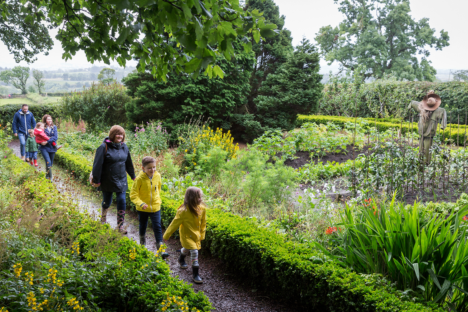 Two families walking along a trail in a garden with a scarecrow.