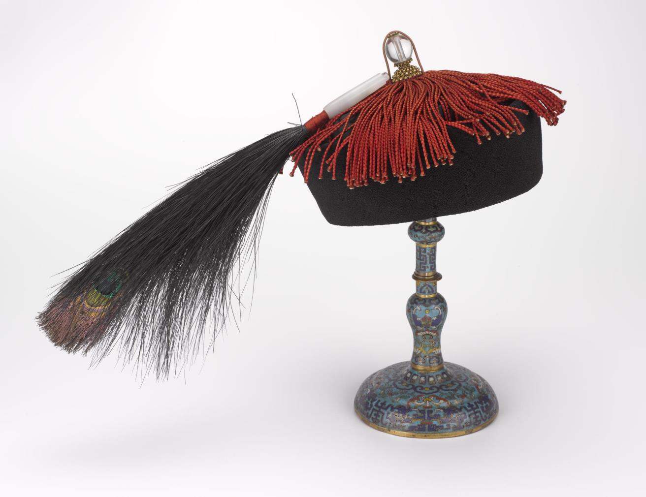 Semi-formal winter hat (jiguan), blue with red tassels and a plume, and with a glass button on top indicating fifth rank official: China, Qing Dynasty, 19th century AD. Lent by Her Majesty the Queen.