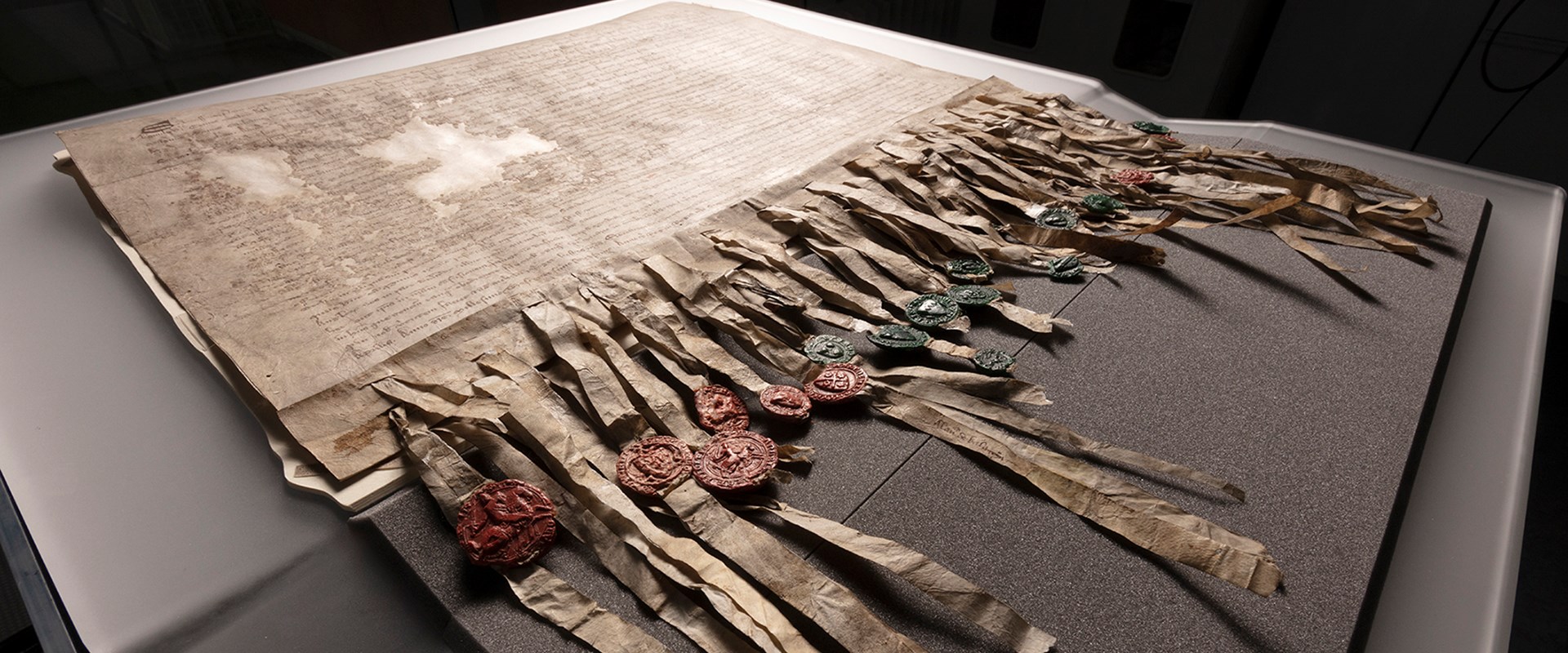 The Declaration of Arbroath laid out on a grey foam protective base set against a black background. The Declaration has dense Latin script across it with two small patches missing, and many strips dangling from the bottom affixed with green and red wax seals.