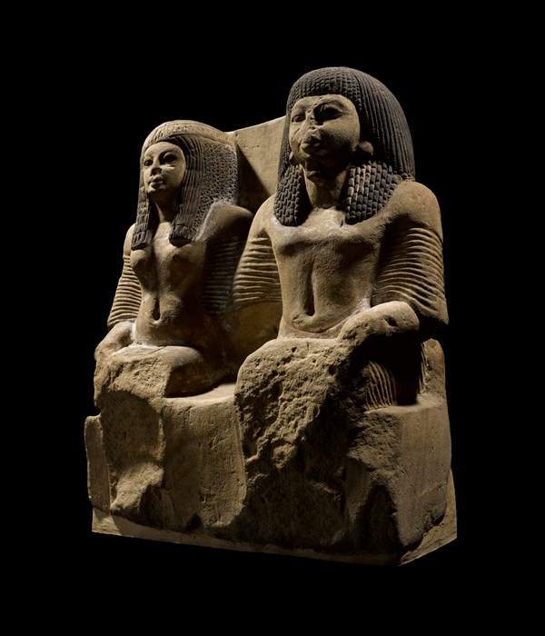Pair statue in fine yellow sandstone of a Chief of the Medjai (police) and his wife seated side by side, wearing long pleated robes, collars and heavy wigs.