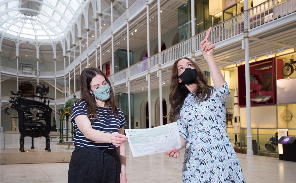 Two women in facemasks hold a museum map, one of them is pointing up towards something.