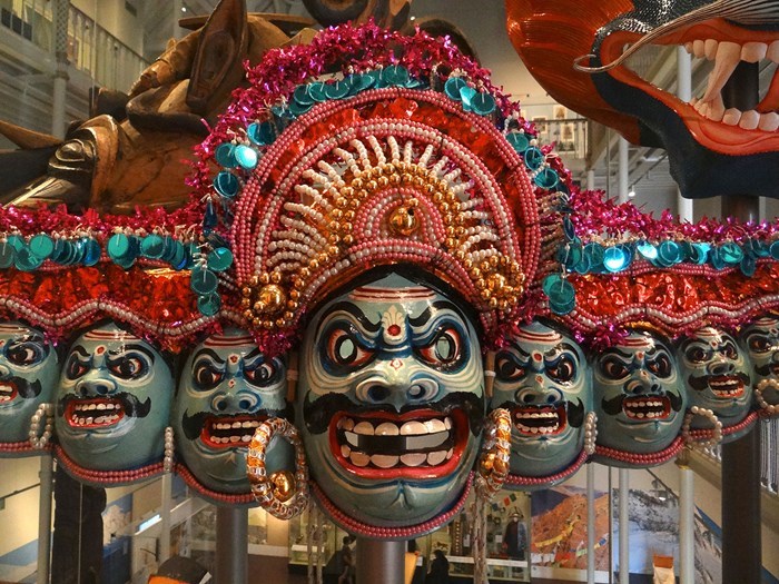 A colourful mask of many blue faces painted with exaggerated eyebrows, teeth, and moustaches under a sparkling headdress of pink, red, blue, pearl, and gold.