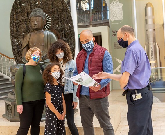 A family stands together with a museum staff member in the grand gallery. They are all wearing masks and looking at a map.