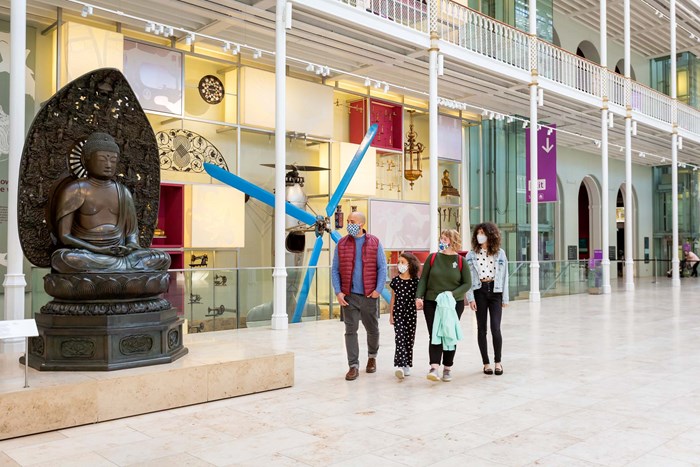 A family stand in the large open space of the Grand Gallery looking at a Buddha statue. They are all wearing masks.