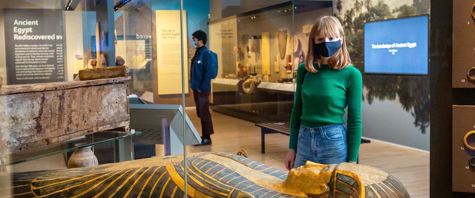 Two visitors wearing masks look at Ancient Egyptian items in glass cases.
