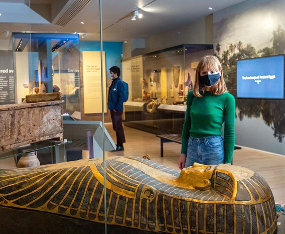 Two visitors wearing masks look at Ancient Egyptian items in glass cases.