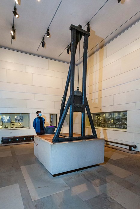 Man in blue shirt and face mask stands behind a guillotine in the Scottish History and Archaeology galleries.