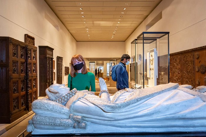 A visitor wearing a mask looks at a replica Mary Queen of Scots' tomb in the foreground. It is a statue of the queen lying down with her hands folded in prayer.