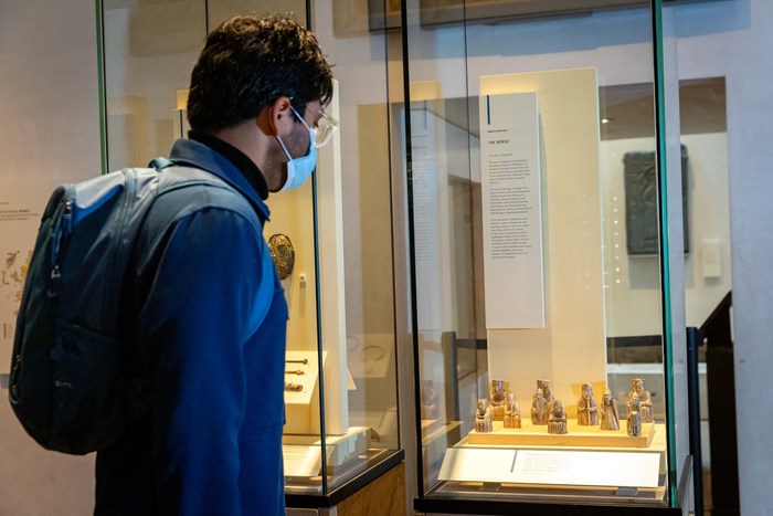 Man in a blue shirt with rucksack and face mask stands in front of the Lewis chess pieces in the Scottish History galleries