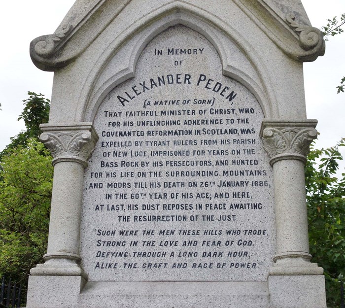 Memorial of Alexander Peden. Large white stone with craved pillars and a long inscription in black with trees in the background.