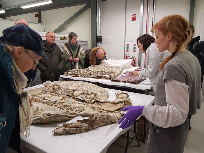 A conservator wearing latex gloves shows some textiles laid out on tables to visitors on a tour