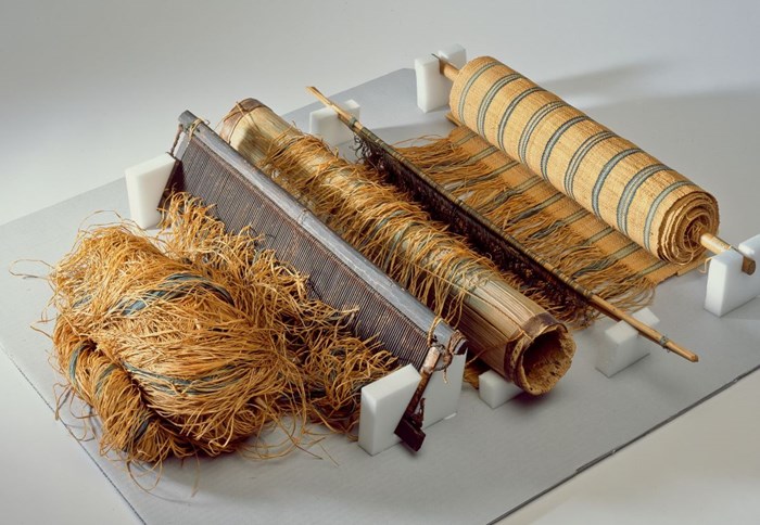 A small loom propped dup by several white blocks. Rough barkcloth strands on the left pass over a roller and comb-like barrier to emerge finely woven on the right.