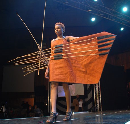 Woman wearing bright orange barkcloth walks down a runway with her left arm held out. Long wooden sticks are across her torso couched in black pouches on the outift.
