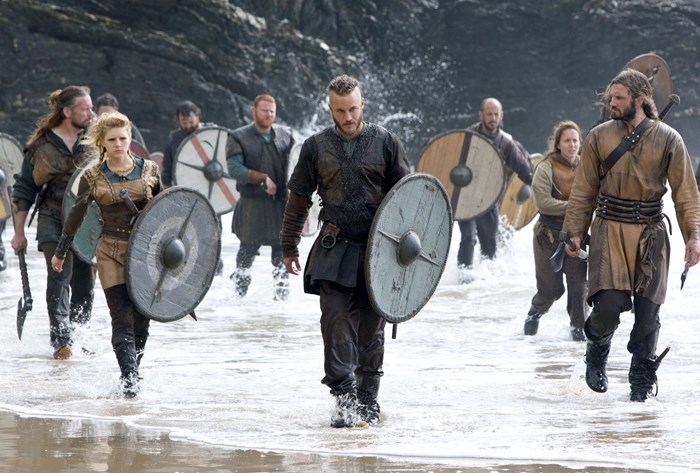 Still from the TV show Vikings, showing the actors in Viking costume and carrying round shields marching in the surf on a beach.