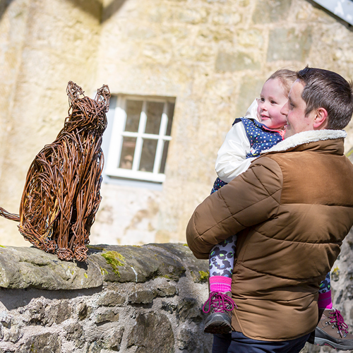 Colour photo of a man holding a small child whilst looking at a willow sculpture of a cat sitting on a wall.