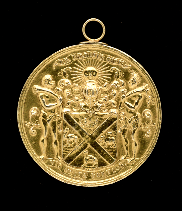 Shiny gold medal that includes the figures of an Indigenous American and an African man. The Latin motto translates to ‘Where the world expands, its united strength is stronger'.