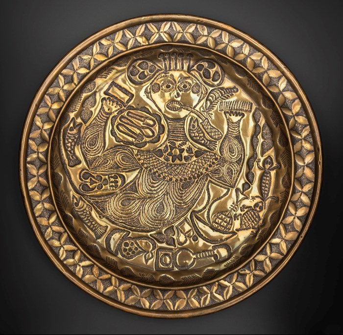 Circular brass embossed tray featuring a female-presenting figure with raised hands, probably Mami Wata, surrounded by fish and lizard and cross patterned outer border.