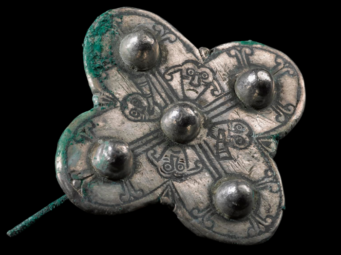 Silver brooch shaped like a four-leaf clover with four faces surrounding a central nub. Two blow horns, the others' ears ring