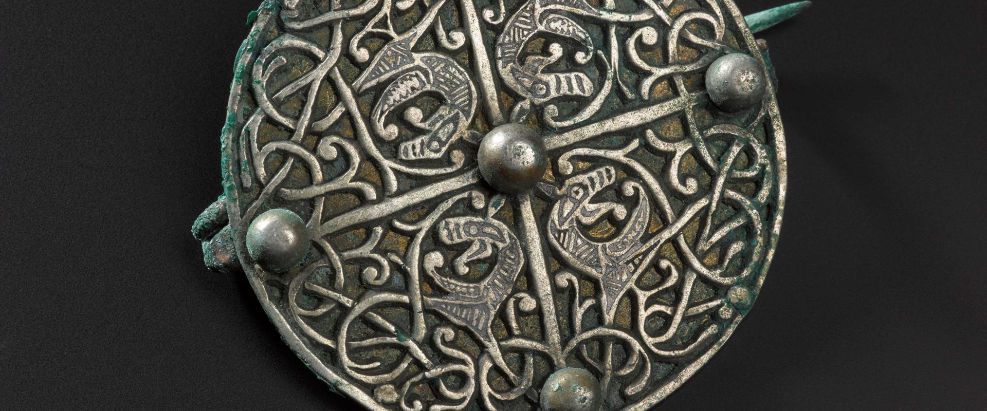 Circular metal brooch on a black background decorated with vine-like patterns and four mysterious beasts in the centre.
