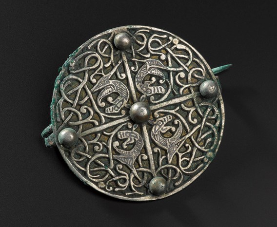 Circular metal brooch on a black background decorated with vine-like patterns and four mysterious beasts in the centre.