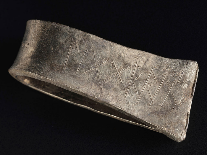 A flattened silver arm-ring resembling a hollow axe head with finely carved runes across its surface