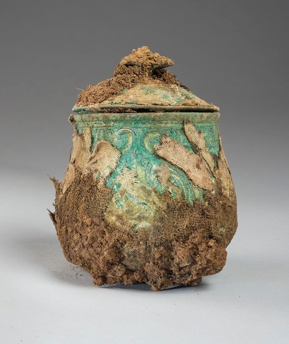Pear-shaped vessel, corroded green with fraying brown textile clinging to it in patches. Naturalistic patterns are hinted at beneath.