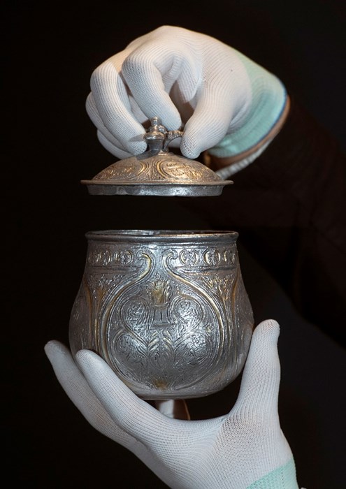 A 3D reproduction of the vessel is held up by two hands in white gloves. One hand gently lifts the vessel's lid several inches up.