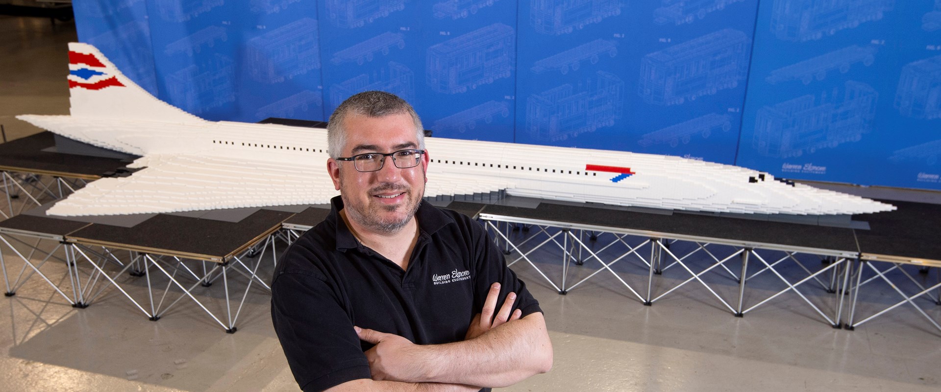 A person with there arms folded standing in front of a lego model of a concorde plane.