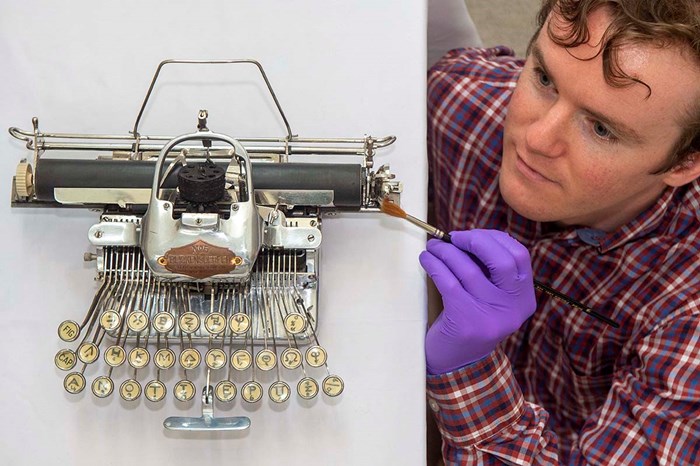 PhD student James Inglis brushing a Blickensderfer featherweight typewriter with a paintbrush